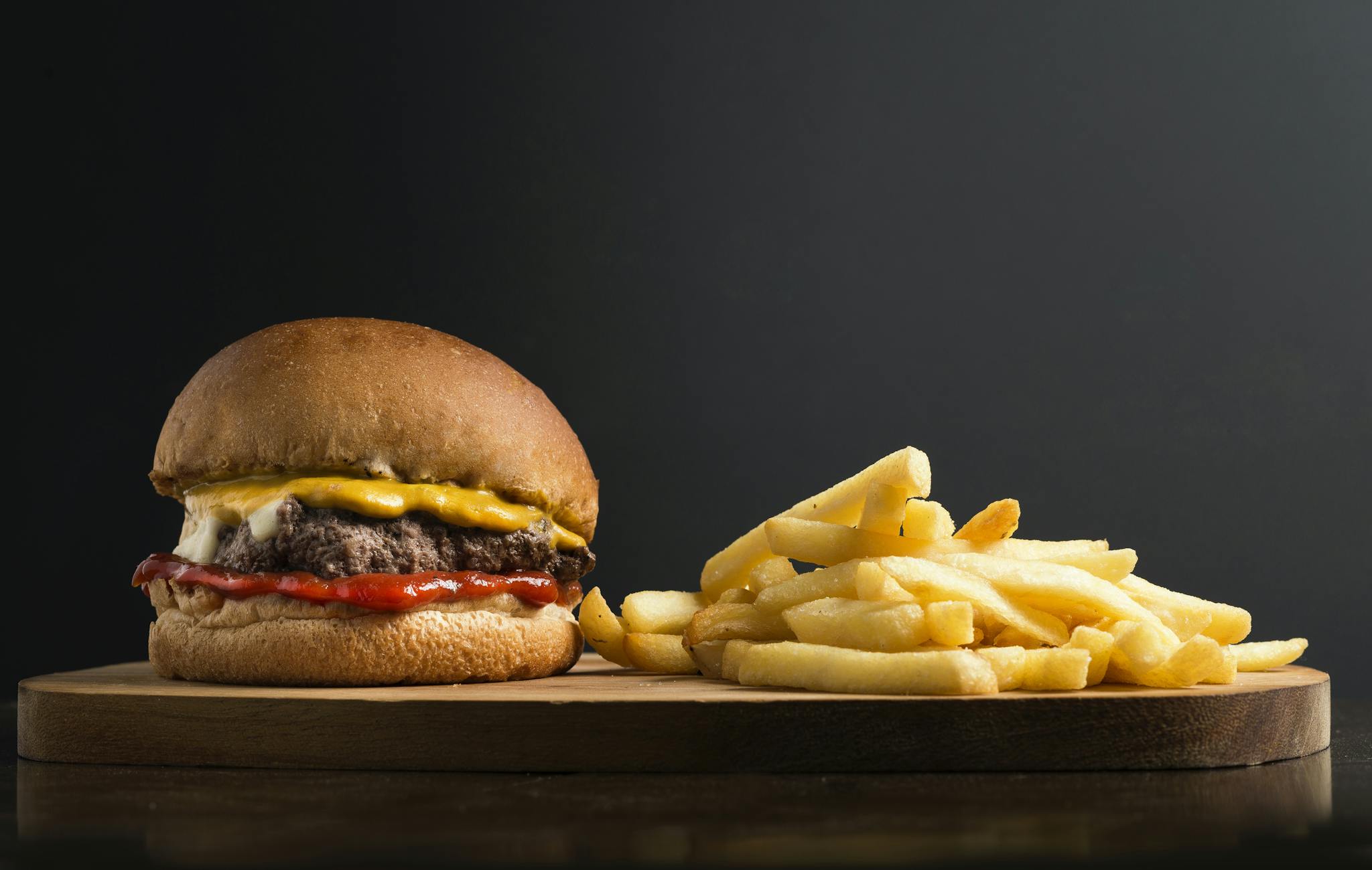 Appetizing burger with meat patty ketchup and cheese placed on wooden table with crispy french fries against black background